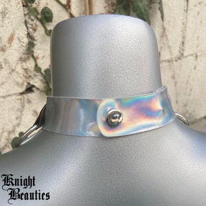 May include: A holographic choker necklace with a silver snap closure and metal ring attachments. The iridescent collar is displayed on a mannequin. The text "Knight Beauties" is printed at the bottom.