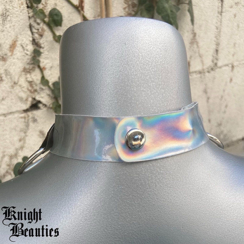 May include: A holographic choker necklace with a silver snap closure and metal ring attachments. The iridescent collar is displayed on a mannequin. The text "Knight Beauties" is printed at the bottom.