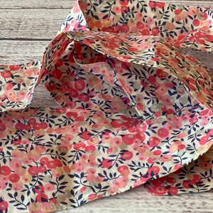 Puede incluir: Una diadema de tela larga y fina con estampado floral con flores rosas y blancas sobre un fondo crema. La diadema est&aacute; doblada por la mitad y tiene un sutil patr&oacute;n azul.