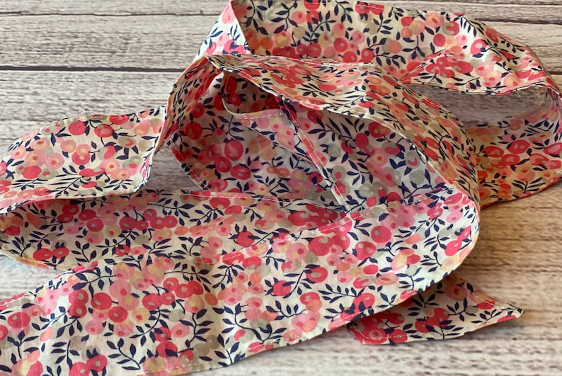 Puede incluir: Una diadema de tela larga y fina con estampado floral con flores rosas y blancas sobre un fondo crema. La diadema est&aacute; doblada por la mitad y tiene un sutil patr&oacute;n azul.