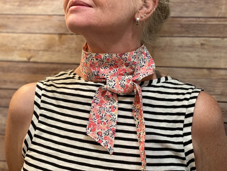 Puede incluir: Un pa&ntilde;uelo de cuello con estampado floral blanco y rosa atado alrededor del cuello de una persona. El pa&ntilde;uelo est&aacute; atado en un lazo en la parte delantera.