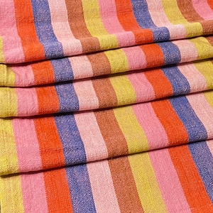 May include: Close-up of folded fabric with vertical stripes in shades of pink, orange, yellow, brown, and blue. The textile has a woven texture, and the stripes vary in width. The fabric is layered, showing the pattern repeating.