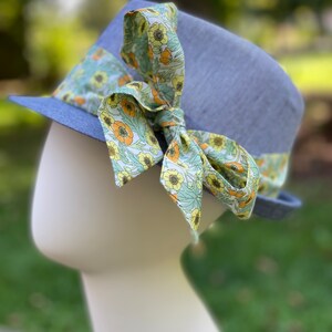 Puede incluir: Un sombrero fedora azul claro con una cinta de estampado floral atada en un lazo. La cinta es verde, amarilla y naranja.