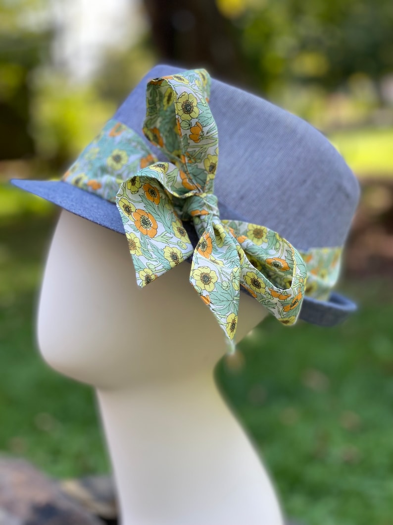 Puede incluir: Un sombrero fedora azul claro con una cinta de estampado floral atada en un lazo. La cinta es verde, amarilla y naranja.