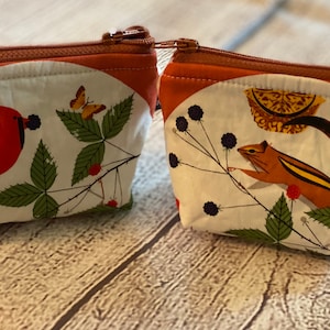 May include: Two small zippered pouches with a white background and a nature-themed design. One pouch features a red cardinal bird with a butterfly, while the other features a brown squirrel with a lemon slice. Both pouches have a brown zipper and a brown top flap.