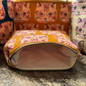 May include: Several cat-patterned cosmetic bags in different colours. The bags display a repeating design of a cat's face in orange, pink, and purple hues. One bag is open, showing a light pink interior and a gold zip.