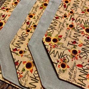 May include: Four placemats with a beige background and a pattern of sunflowers, leaves, and the word "Thanks" repeated. The placemats have a gray linen border.
