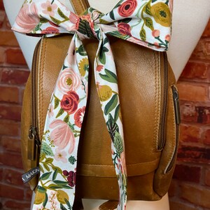 Puede incluir: Una mochila de cuero marr&oacute;n con un lazo de tela con estampado floral atado alrededor de las correas. La tela tiene flores rosas, rojas y verdes sobre un fondo blanco. La mochila tiene una cremallera y un peque&ntilde;o bolsillo en la parte delantera. El nombre de la marca "Makin" es visible en el bolsillo delantero.
