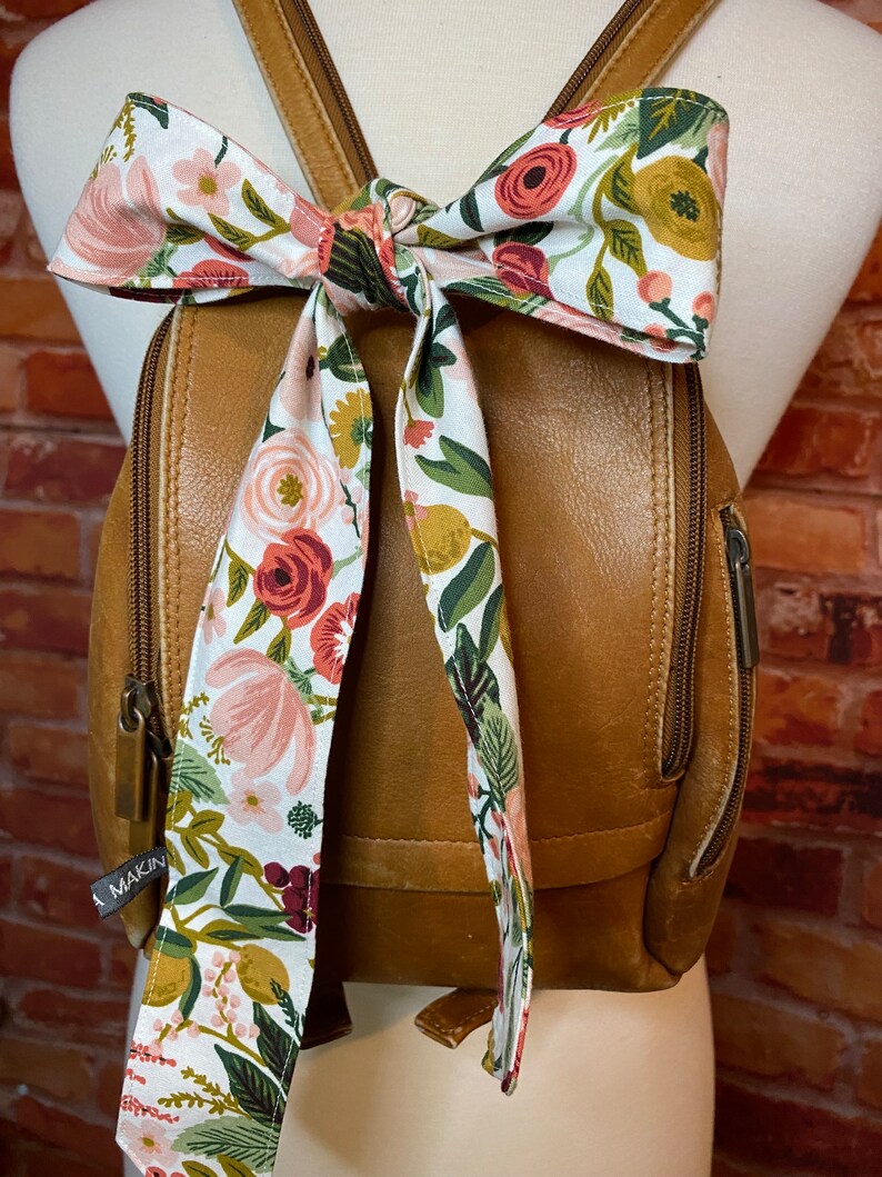 Puede incluir: Una mochila de cuero marr&oacute;n con un lazo de tela con estampado floral atado alrededor de las correas. La tela tiene flores rosas, rojas y verdes sobre un fondo blanco. La mochila tiene una cremallera y un peque&ntilde;o bolsillo en la parte delantera. El nombre de la marca "Makin" es visible en el bolsillo delantero.