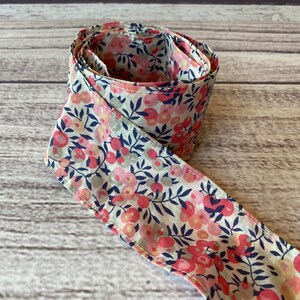 Puede incluir: Una cinta de tela blanca con un estampado floral rosa y azul. La cinta est&aacute; enrollada y anudada.