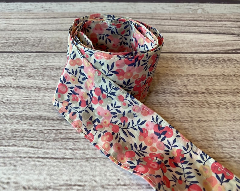 Puede incluir: Una cinta de tela blanca con un estampado floral rosa y azul. La cinta est&aacute; enrollada y anudada.