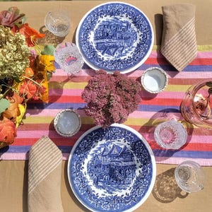 Może przedstawiać: Nakrycie stołu ze stolnicą w paski, dwoma talerzami w niebiesko-białe wzory i bukietem różowych kwiatów w środku. Na stole znajdują się również różne szklanki i małe miski.