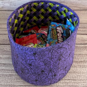 Op de afbeelding: Een paarse stoffen Halloween-snoepemmer met een spinnenweb- en spinnenontwerp. De emmer is gevuld met verschillende snoepzakjes, waaronder Sour Patch Kids, Sour Bite Crawlers en Trolli Sour Brite Crawlers.
