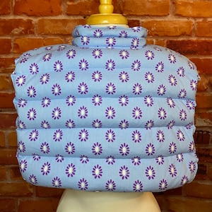 May include: A light blue quilted vest with a white and purple floral pattern. The vest has a high collar and is made of a soft, comfortable fabric.