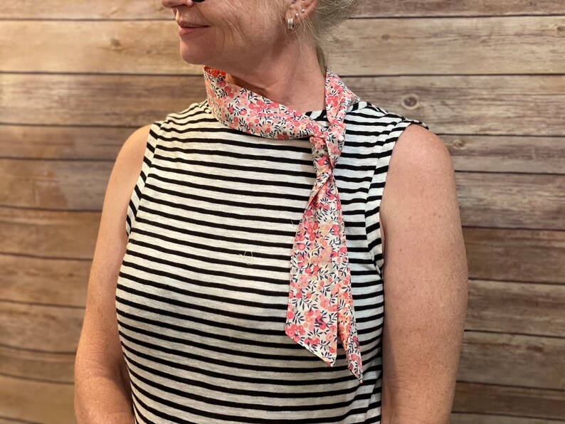 Puede incluir: Una mujer con una camisa sin mangas a rayas con una bufanda de estampado floral rosa y blanco atada alrededor del cuello.