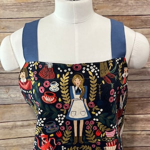 May include: A blue and black apron with a whimsical print featuring Alice in Wonderland characters, teacups, and flowers. The apron has adjustable straps.