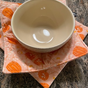 以下が含まれることがあります： わずかにテクスチャ加工された縁のある白いセラミックボウルが、ピンクとオレンジの南瓜模様の鍋敷きの上に置かれています。鍋敷きは白い背景にオレンジの南瓜と緑のつるが描かれています。