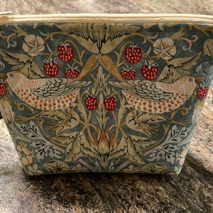 May include: A small, rectangular zippered pouch with a botanical print featuring birds, strawberries, and foliage in shades of blue, beige, and red. The zipper is beige, and a small black tag is attached to the side.