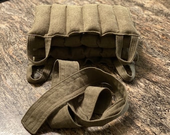 Puede incluir: Una almohadilla térmica rectangular de color verde oliva con múltiples secciones segmentadas. La almohadilla tiene dos asas de bucle a cada lado y una correa larga a juego. La tela parece ser un material suave y texturizado.