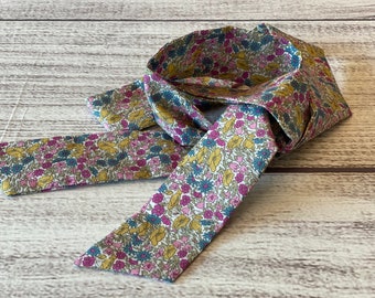 May include: A floral patterned fabric necktie in shades of pink, yellow, blue, and green. The tie is made of a soft fabric and is tied in a loose knot. The tie is laying on a wooden surface.