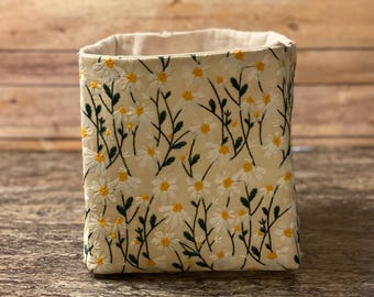 May include: A rectangular fabric storage bin with a cream-colored background and embroidered white daisies with yellow centers and green stems. The interior is a light beige color. The bin is sitting on a brown surface.