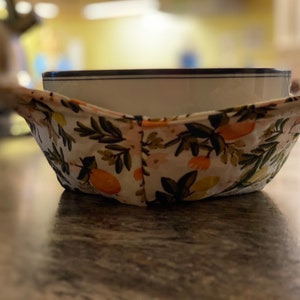 May include: A white ceramic bowl with a blue rim sits inside a floral patterned bowl cozy. The cozy is made of fabric with a white background and features a green leafy pattern with orange citrus fruit.
