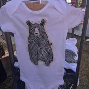 May include: White baby onesie with a black and white bear graphic. The onesie has the word "carters" printed on the tag.