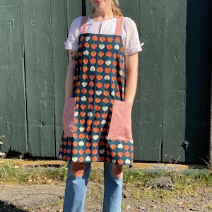 May include: A woman wearing a blue and white apron with a strawberry print. The apron has two large pockets with red fabric lining. She is wearing blue jeans and brown clogs.
