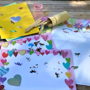 May include: A selection of colourful stationery, including sheets of paper in blue, green, and yellow, decorated with hearts, unicorns, and horse-shaped confetti. Pencils are in a cylindrical container. A clear glass is also present.