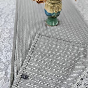 Op de afbeelding: Een grijze en witte gestreepte tafelrunner met een wit geborduurd ontwerp. De tafelrunner heeft een label met de tekst "MAIDS F".