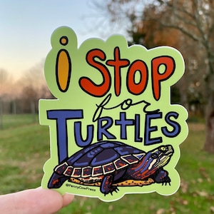 May include: A die-cut sticker with the text "i Stop for Turtles" in orange, blue, and black. The sticker features a detailed illustration of a turtle with a dark shell and red and orange markings. The background is a light green.