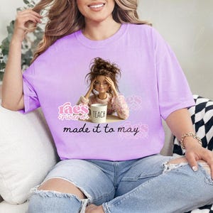 May include: A light purple t-shirt featuring a graphic of a doll with the word "TEACH" on a mug. The text "rae's riviera" and "made it to may" are also on the shirt. The shirt is worn with ripped blue jeans.