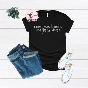 May include: Black t-shirt with the text "CHRISTMAS TREES and Jesus please" in white script. The shirt is paired with blue jeans and white canvas sneakers. A sprig of greenery is in the upper left corner.