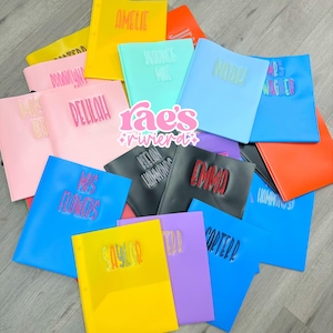 May include: A collection of colorful, personalized binders in various shades, including yellow, pink, blue, and orange. Each binder features a name in a contrasting color, such as "Amelie" or "Noah". The binders are arranged on a light gray surface.