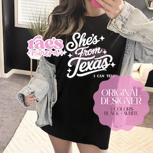 May include: Black t-shirt with the text "She's From Texas" in white script. The shirt also features the text "I Can Tell" and the words "Rae's Riviera" in pink. The design is labeled as the "Original Designer".
