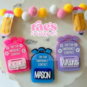 May include: A display of personalized backpack-shaped name tags in pink, blue, and purple, each with a name and the text "TAP FOR EMERGENCY CONTACT." A felt ball garland and pencil-shaped decorations complete the design.