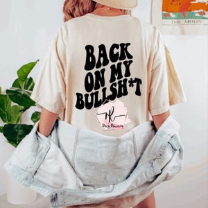 May include: Cream-colored t-shirt with the text "BACK ON MY BULLSH*T" in bold, black letters. The shirt has a relaxed fit and is worn with a light-wash denim jacket. The design includes a logo at the bottom.