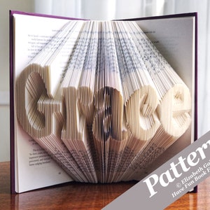 Puede incluir: Un libro doblado en forma de la palabra "Grace". El libro está abierto y las páginas están dobladas para crear las letras. El libro está sobre una mesa de madera. El texto "Pattern" es visible en la esquina inferior derecha de la imagen.