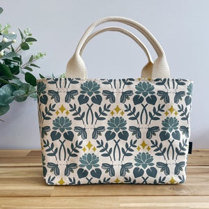 May include: A small, cream-colored tote bag with a floral pattern. The pattern features green leaves, blue flowers, and yellow stars. The bag has two cream-colored handles.