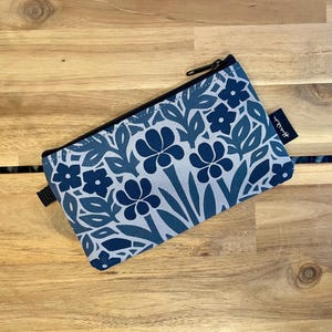 May include: A rectangular zippered pouch featuring a repeating floral pattern in shades of navy blue, medium blue, and light gray. The pouch has a black zipper closure along the top edge and a small black fabric tag with white text on the side.