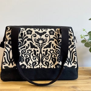 May include: Black and cream tote bag featuring a decorative floral and bat design. The bag has black handles and a black base. The rectangular bag has a zip closure.