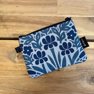 May include: A rectangular zippered pouch featuring a repeating floral pattern in shades of navy blue and light blue on a light gray background. The pouch has a black zipper closure along the top edge and a small black fabric tag with white text.