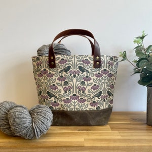 May include: A beige canvas tote bag with brown leather handles and a dark brown bottom. The bag features a repeating pattern of purple flowers, green leaves, black birds, and brown mushrooms. The bag is filled with gray yarn.