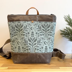 May include: A brown and blue canvas backpack with a dragonfly and floral pattern. The backpack has a brown leather handle and adjustable straps.