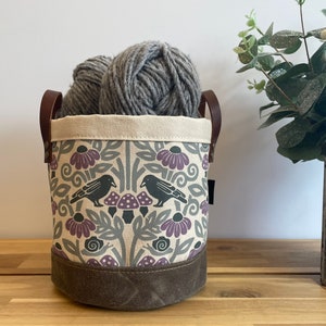 May include: A canvas storage bin with brown leather handles, featuring a floral pattern with purple flowers, green leaves, and black birds. The bin is filled with gray yarn.