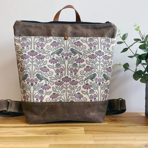 May include: A brown canvas backpack with a floral pattern featuring purple flowers, gray birds, and white mushrooms. The backpack has a brown leather handle and a zipper closure.