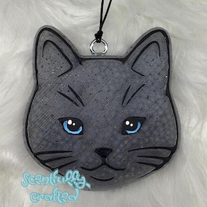 May include: A gray cat-shaped hanging ornament with bright blue eyes and black etched details. The surface has a textured, speckled finish. A black string is attached to a silver metal ring at the top. The text Scentfully Crafted is in light blue.