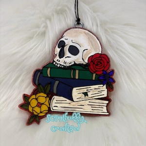 Könnte beinhalten: Ein dekorativer Lufterfrischer mit einem Totenkopf auf einem Stapel Bücher, mit roten und gelben Rosen. Der Text "Scentfully Crafted" ist enthalten. Der Lufterfrischer wird von einer schwarzen Schnur gehalten.