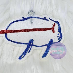 Può includere: Un ornamento glitterato con il logo dei Buffalo Bills. Il logo è bianco con una striscia rossa e un contorno blu, e un anello d'argento per appenderlo. L'ornamento è su uno sfondo bianco e soffice.