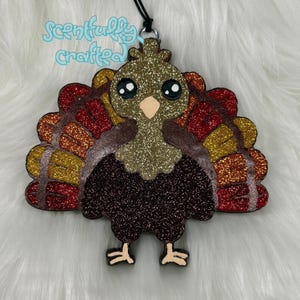 May include: Glittery turkey-shaped ornament with a black string for hanging. The turkey features a brown body, a gold head, and a colorful fan of red, orange, and gold feathers. The text "Scentfully Crafted" is written in light blue.